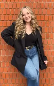 Woman with blonde curly hair stood with one hand on her hip. Wearing black top, black blazer and blue jeans. Stood against a brick wall. Victoria Steele, Marketing Consultant