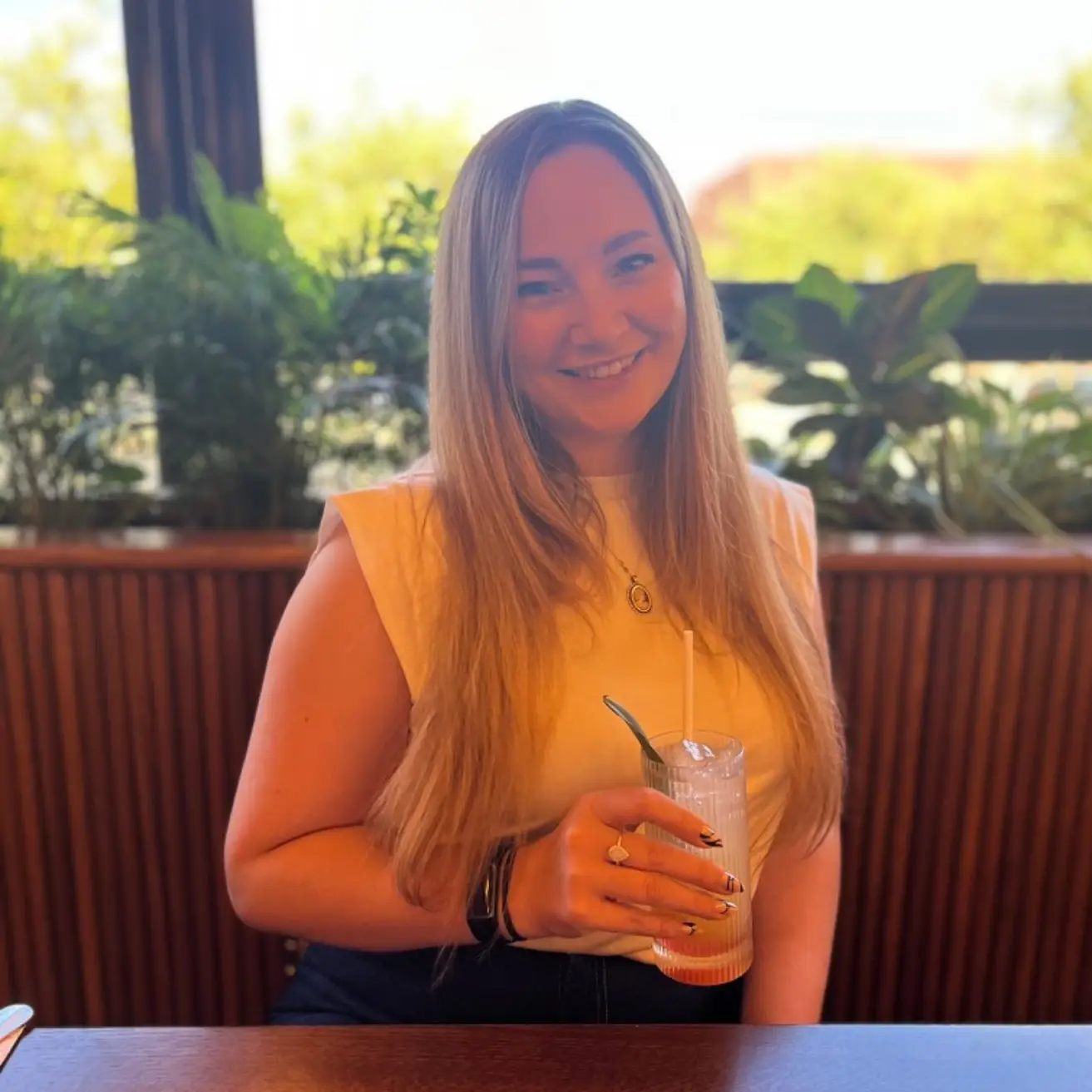 Long brown haired woman sat in front of a window. Wearing a white t-shirt and with a lemonade in front of her. Victoria Steele, Marketing Consultant