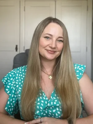 A 35 year old white woman with long brown hair, with arms resting on a desk. Wearing a green dress with white spots. Victoria Steele, Marketing Consultant in the Midlands.