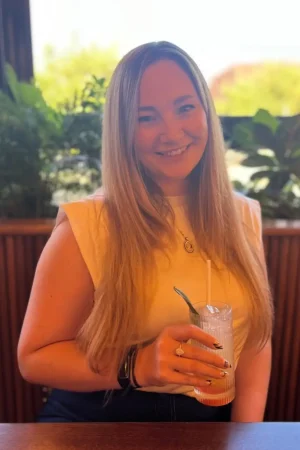 Victoria Steele Long brown haired woman sat in front of a window. Wearing a white t-shirt and with a lemonade in front of her. Victoria Steele, Marketing Consultant